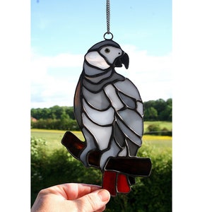 May include: A stained glass grey parrot perched on a branch with red accents. The parrot is hanging from a silver chain.