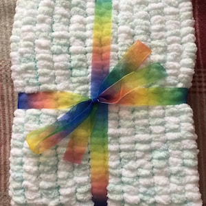 Peut inclure: Une couverture pour bébé en crochet blanc et bleu clair avec un ruban arc-en-ciel noué autour.