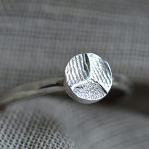 May include: A silver ring with a textured circular design. The ring is made of metal and has a simple band.