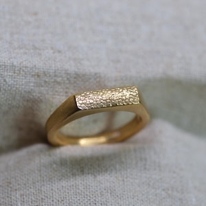 May include: A gold-colored ring with a rectangular textured top. The ring has a smooth, polished band and a textured surface on the top. The ring is set against a neutral-colored fabric background.