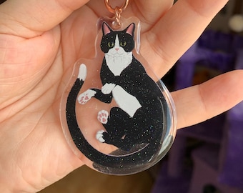 May include: A black and white cat with green eyes is sitting in a clear resin keychain. The cat is curled up with its tail wrapped around its body. The keychain has a sparkly finish.