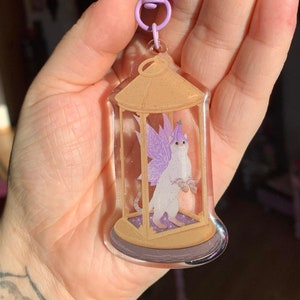 Könnte beinhalten: Ein durchsichtiger Acryl-Schlüsselanhänger mit einer weißen Katze mit violetten Flügeln in einem goldenen Käfig. Der Schlüsselanhänger hat einen violetten Clip.