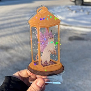 Könnte beinhalten: Eine weiße Katze mit rosa Flügeln und einem kleinen grünen Hut befindet sich in einer durchsichtigen Acryllanterne mit goldenem Rahmen. Die Laterne hat einen goldenen Sockel und ist mit schimmerndem Glitzer verziert.