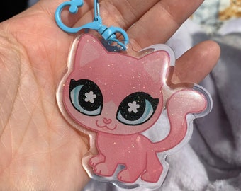 Op de afbeelding: Roze glitterende katten sleutelhanger met grote blauwe ogen en witte bloemaccenten. De sleutelhanger is bevestigd aan een blauwe metalen ring.