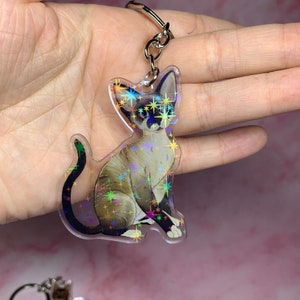 May include: A clear iridescent keychain featuring a cat with a white and brown coat. The cat is sitting with its tail curled around its legs. The keychain is decorated with a holographic glitter effect.