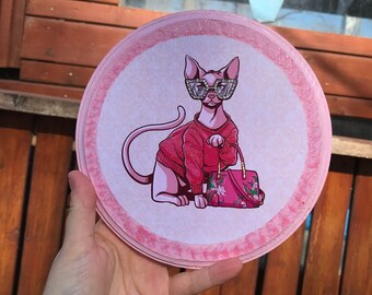 Puede incluir: Un posavasos redondo rosa y blanco con una imagen de dibujos animados de un gato sin pelo con gafas de sol y un suéter rojo que sostiene un bolso de mano rosa y floral.