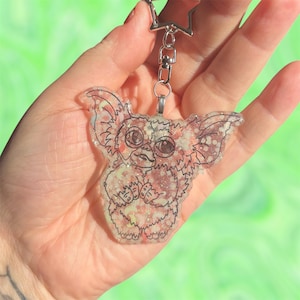 May include: A clear keychain featuring a cartoon creature with large eyes, ears, and a fluffy body. The creature is outlined in black and filled with glittery pink and white accents. The keychain has a silver star-shaped clasp.