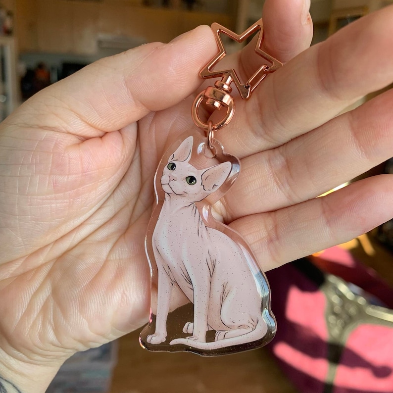 May include: A pink and white hairless cat keychain charm with a gold star charm attached to a silver ring. The keychain is made of clear acrylic.