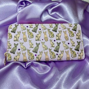 Könnte beinhalten: Eine weiße Geldbörse mit einem sich wiederholenden Muster von Sphinx-Katzen in verschiedenen Posen. Die Katzen sind eine Mischung aus hell- und dunkelgrau mit violetten Strasssteinen, die über das gesamte Design verteilt sind.