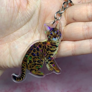 Peut inclure: Un porte-clés en acrylique transparent représentant un chat Bengal brun et noir avec des accents de paillettes irisées. Le chat est assis, la tête tournée sur le côté et les yeux fixés sur le spectateur.
