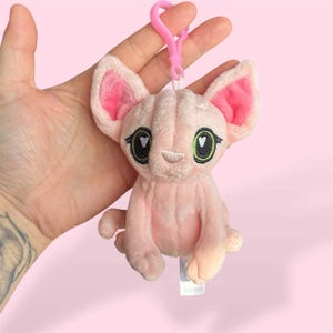 May include: A pink Sphynx cat plush keychain with large ears and green eyes. The cat has a pink nose and a pink clip attached to its head. The keychain is held against a pink background.