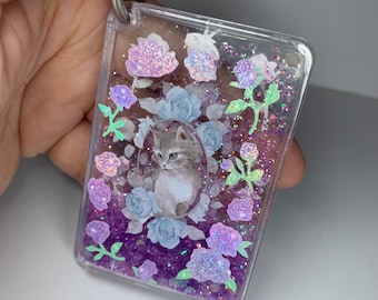 Peut inclure: Un porte-clés transparent avec un fond de paillettes violettes et un petit chaton entouré de fleurs. Les fleurs sont blanches, roses et violettes.