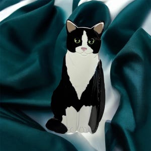 May include: Black and white cat brooch with green eyes. The cat is sitting with its front paws together and its back paws slightly apart. The brooch is made of metal and has a shiny finish.