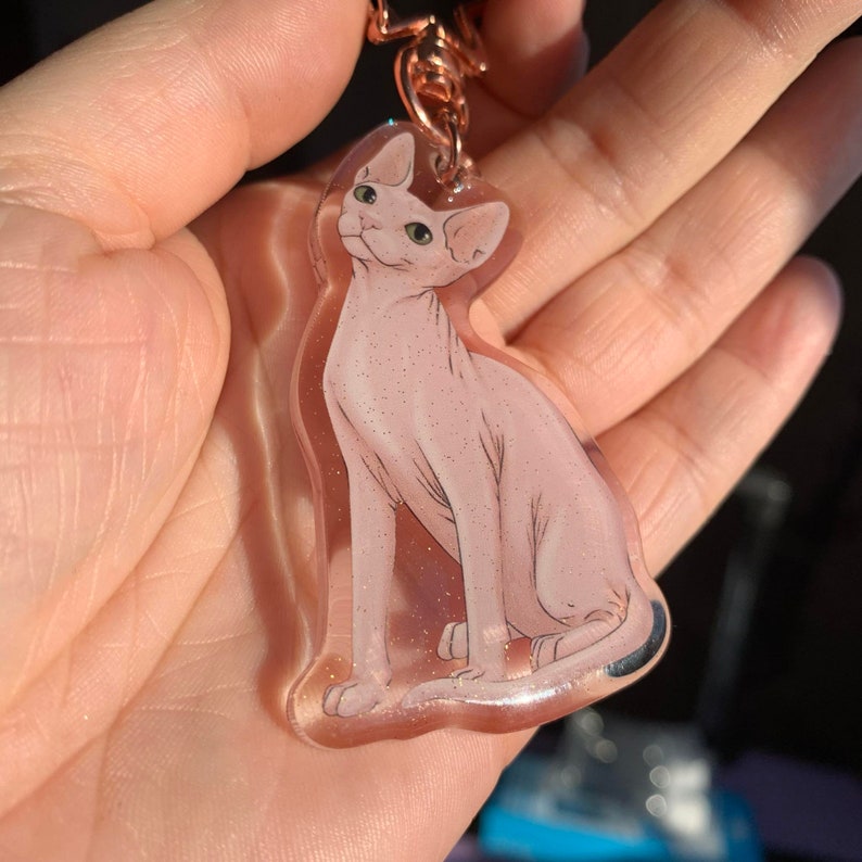 May include: A clear pink and white resin keychain featuring a detailed illustration of a hairless cat. The cat is sitting with its tail curled around its legs. The keychain has a gold metal ring.
