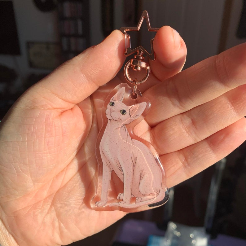 May include: A clear acrylic keychain featuring a pink hairless cat with green eyes. The keychain is attached to a rose gold star-shaped charm and a rose gold chain.