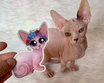 以下が含まれることがあります： 花冠と大きな青い目をしたピンクの猫がステッカーに描かれています。ステッカーは手に持たれており、背景には毛のない本物の猫がいます。