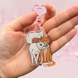 Puede incluir: Un llavero de acrílico transparente con un cierre rosa en forma de corazón y una ilustración de dos gatos. Un gato es blanco con un collar dorado y el otro es naranja con una pajarita verde. Los gatos están juntos con sus colas entrelazadas.