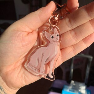May include: A clear keychain with a pink hairless cat sitting on a white background. The keychain is attached to a rose gold star-shaped charm.