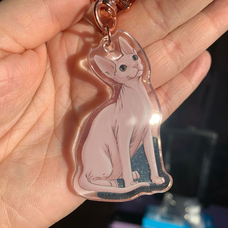 May include: A clear keychain with a pink and white Sphynx cat illustration. The cat is sitting with its head tilted up and its eyes open.