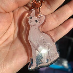 May include: A clear keychain with a pink and white cat with green eyes. The cat is sitting and has glitter embedded in the resin.