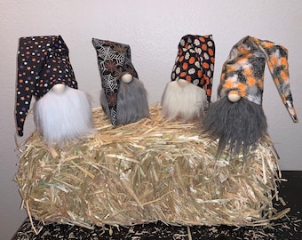 May include: Four Halloween gnomes with different patterns on their hats. The gnomes are sitting on a bed of straw.