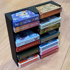 May include: A black playing card holder, stacked with multiple decks of cards. The decks display vibrant designs, including Disney's "Cats & Dogs" and "Stitch" themes, and the "Bicycle" brand. The decks are labeled "Air-Cushion Finish" and "Made in USA".