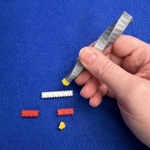 Könnte beinhalten: Eine Hand hält eine grau-gelbe Pinzette und hebt einen gelben Baustein auf. Mehrere rote, weiße und gelbe Bausteine ​​sind auf einer blauen Oberfläche verstreut. Die Pinzette hat ein geometrisches Design.