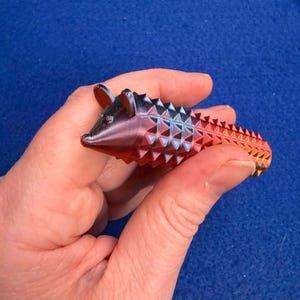 May include: A small, colourful, 3D-printed armadillo figurine. The armadillo is primarily red, with gradients of blue, purple, and yellow. It has a textured, spiky shell and small ears. The figurine is held in a hand against a blue background.