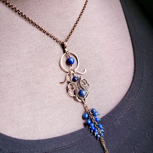 K&ouml;nnte beinhalten: Eine handgefertigte Halskette mit einer Bronzekette und einem Anh&auml;nger. Der Anh&auml;nger hat ein filigranes Design mit tiefblauen Perlen und einer Ansammlung kleinerer Perlen am unteren Ende. Die Halskette wird vor einem neutralen Hintergrund gezeigt.