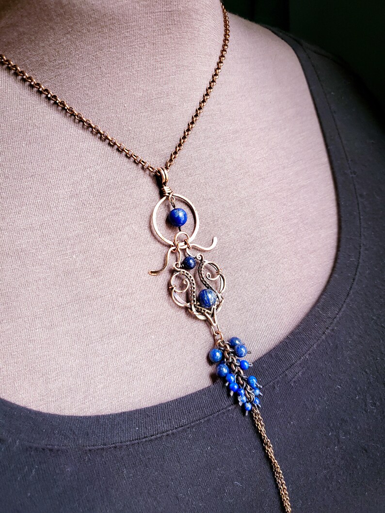 K&ouml;nnte beinhalten: Eine handgefertigte Halskette mit einer Bronzekette und einem Anh&auml;nger. Der Anh&auml;nger hat ein filigranes Design mit tiefblauen Perlen und einer Ansammlung kleinerer Perlen am unteren Ende. Die Halskette wird vor einem neutralen Hintergrund gezeigt.