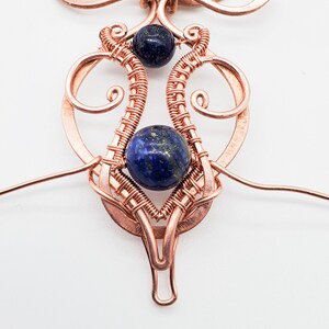 K&ouml;nnte beinhalten: Ein handgefertigter Kupferdraht-Anh&auml;nger mit zwei dunkelblauen Lapis-Lazuli-Edelsteinperlen. Der Anh&auml;nger hat ein kompliziertes, offenes Design mit spiralf&ouml;rmigen Details und einem Drahtk&auml;fig um die untere Perle.