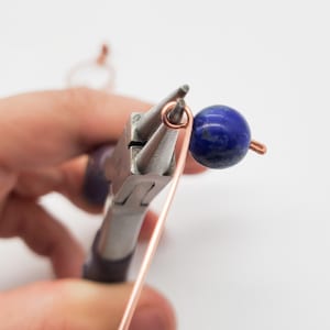 K&ouml;nnte beinhalten: Nahaufnahme der Schmuckherstellung. Kupferdraht wird mit einer Zange zu einer Schlaufe geformt. Eine dunkelblaue, runde Perle ist am Draht befestigt. Der Hintergrund ist wei&szlig;.