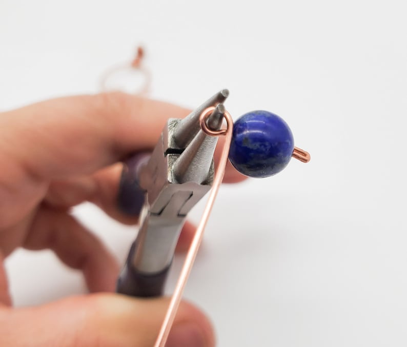 K&ouml;nnte beinhalten: Nahaufnahme der Schmuckherstellung. Kupferdraht wird mit einer Zange zu einer Schlaufe geformt. Eine dunkelblaue, runde Perle ist am Draht befestigt. Der Hintergrund ist wei&szlig;.