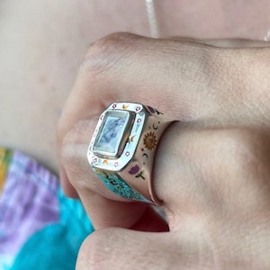 May include: A silver ring with a rectangular, light-colored stone. The ring features celestial-themed designs, including a sun, moon, and stars, with a turquoise band. The ring is worn on a finger.