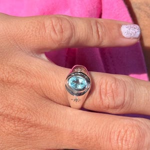 May include: A silver-toned ring featuring a central oval-shaped light blue gemstone set in a smooth metal band. The band is accented with two small, engraved starburst designs positioned above and below the central stone setting.