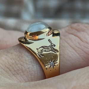 May include: Gold-tone ring featuring a large, round, light-colored stone. The ring's band has a detailed design of a running hare, stars, and a small diamond accent. The ring is a unique piece of jewelry.