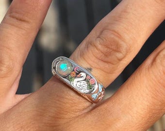 May include: A silver ring featuring a luminous opal stone. The ring's band is adorned with a white swan, colorful flowers, and blue accents. The ring is a piece of jewelry.