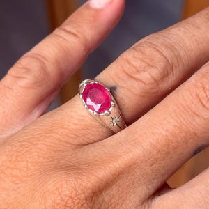 May include: A silver ring featuring a central oval-cut vibrant pinkish-red gemstone held in a scalloped bezel setting. The band is crafted from polished silver and includes a small, star-shaped engraving with a tiny inset diamond accent.