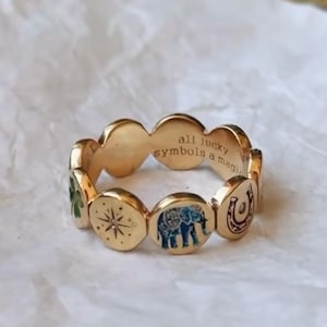 Op de afbeelding: Goudkleurige ring met gelukssymbolen: een klavertjevier, een kompasroos, een olifant en een hoefijzer. De ring heeft de inscriptie "all lucky symbols a magic". De ring ligt op een witte ondergrond.