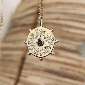 Può includere: Collana d'oro con un ciondolo rotondo che presenta una pietra preziosa blu circondata da cinque conigli in un cerchio. Il ciondolo è inciso con stelle e lune.