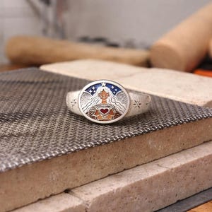 Puede incluir: Un anillo de plata con una cara ovalada que presenta a una mujer con cabello largo, sosteniendo un coraz&oacute;n. El anillo tiene un fondo estrellado azul y blanco. El anillo est&aacute; sobre una superficie met&aacute;lica texturizada.