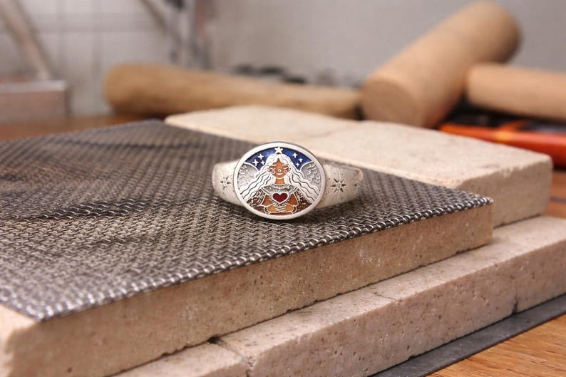 Puede incluir: Un anillo de plata con una cara ovalada que presenta a una mujer con cabello largo, sosteniendo un coraz&oacute;n. El anillo tiene un fondo estrellado azul y blanco. El anillo est&aacute; sobre una superficie met&aacute;lica texturizada.