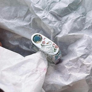 Puede incluir: Un anillo plateado con una parte superior rectangular con una piedra preciosa azul. El anillo está decorado con un cisne, flores y una luna creciente. El anillo está sobre una superficie de papel blanco arrugado.
