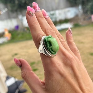 Puede incluir: Un anillo de plata con un diseño de rana verde tallada. La rana está sobre un fondo verde oscuro dentro de un marco ovalado plateado. El anillo se lleva en una mano con uñas decoradas en rosa y blanco.