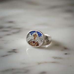 Puede incluir: Un anillo de plata con una cara ovalada con una ilustraci&oacute;n colorida. La ilustraci&oacute;n representa una figura con una corona, alas y un coraz&oacute;n. La banda del anillo tiene un dise&ntilde;o de estrella.