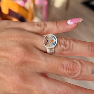 Puede incluir: Anillo de plata con una cara circular que presenta una ilustraci&oacute;n detallada de la cara de una mujer, un coraz&oacute;n y estrellas. El anillo tiene un dise&ntilde;o de estrella en la banda. La cara del anillo tiene detalles en rojo, marr&oacute;n y azul.