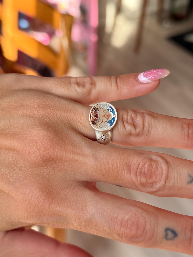 Puede incluir: Anillo de plata con una cara circular que presenta una ilustraci&oacute;n detallada de la cara de una mujer, un coraz&oacute;n y estrellas. El anillo tiene un dise&ntilde;o de estrella en la banda. La cara del anillo tiene detalles en rojo, marr&oacute;n y azul.