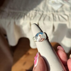 Puede incluir: Anillo de plata con una cara ovalada detallada que presenta una ilustraci&oacute;n colorida de una mujer con un coraz&oacute;n y un dise&ntilde;o de sol. El anillo se muestra en un soporte c&oacute;nico blanco. El texto "THE SUN" es visible en el soporte.