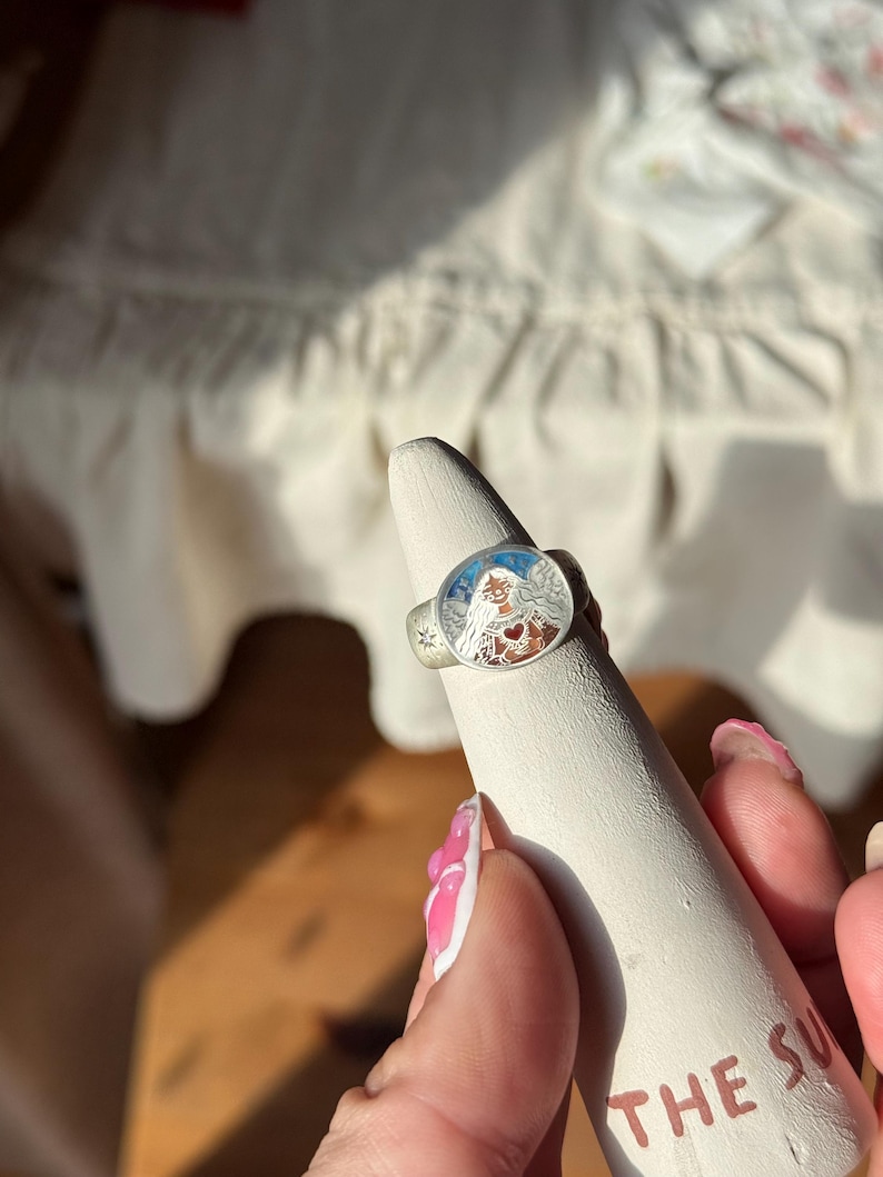 Puede incluir: Anillo de plata con una cara ovalada detallada que presenta una ilustraci&oacute;n colorida de una mujer con un coraz&oacute;n y un dise&ntilde;o de sol. El anillo se muestra en un soporte c&oacute;nico blanco. El texto "THE SUN" es visible en el soporte.