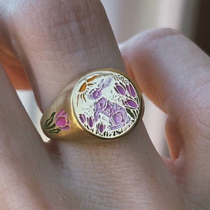 May include: A gold ring with a white enamel design of a rabbit in a field of flowers. The flowers are pink, green, and yellow.